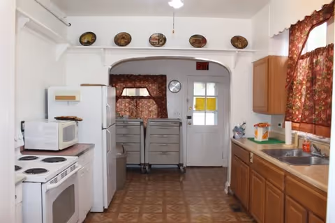 A kitchen with white stove and refrigerator, wooden cabinets and sink under a floral-curtained window, and a door at the far end.