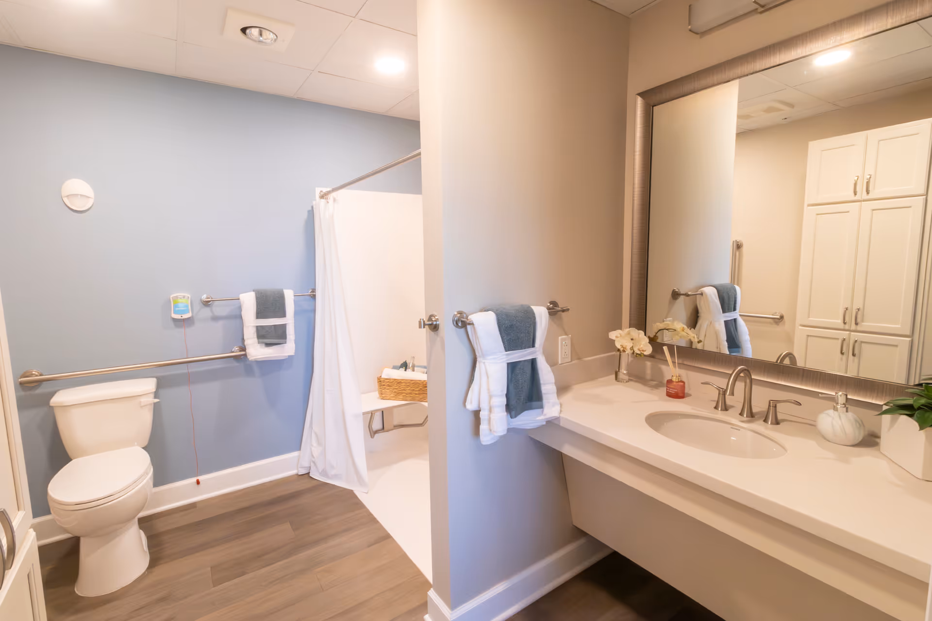 A clean and modern bathroom with a white toilet, a walk-in shower with a white curtain, and a vanity with a sink, mirror, and countertop. The walls are painted light blue and beige, and there are towels hanging on metal bars. The floor has wood-like flooring, and there are some decorative items on the countertop including a small plant and a reed diffuser.