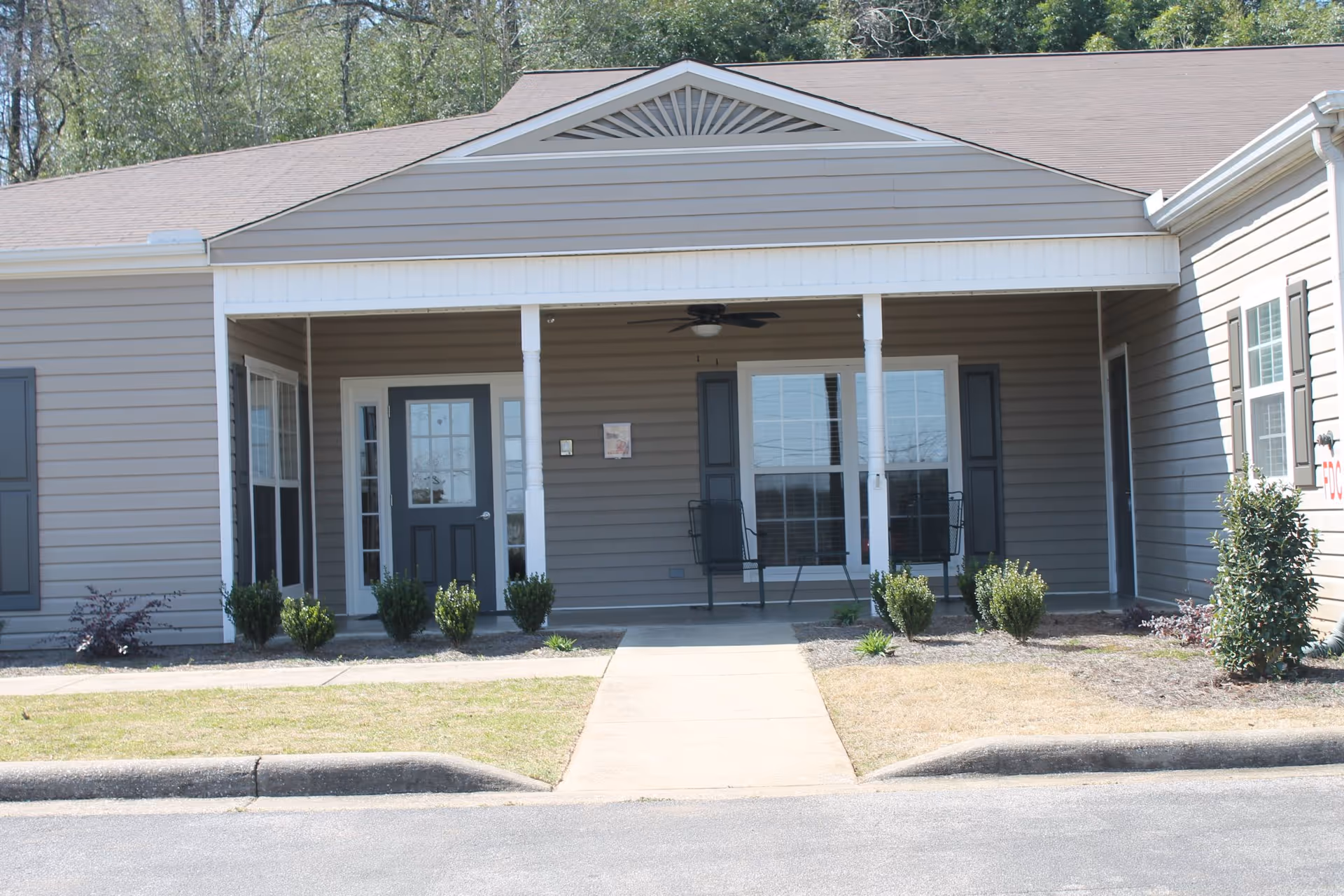 Front exterior view of a single-story building with beige siding and a covered porch area. The porch has two white columns, a ceiling fan, two black chairs, and a black door with glass panels. There are several small bushes and plants in front of the porch, and a concrete walkway leads to the entrance from the street.