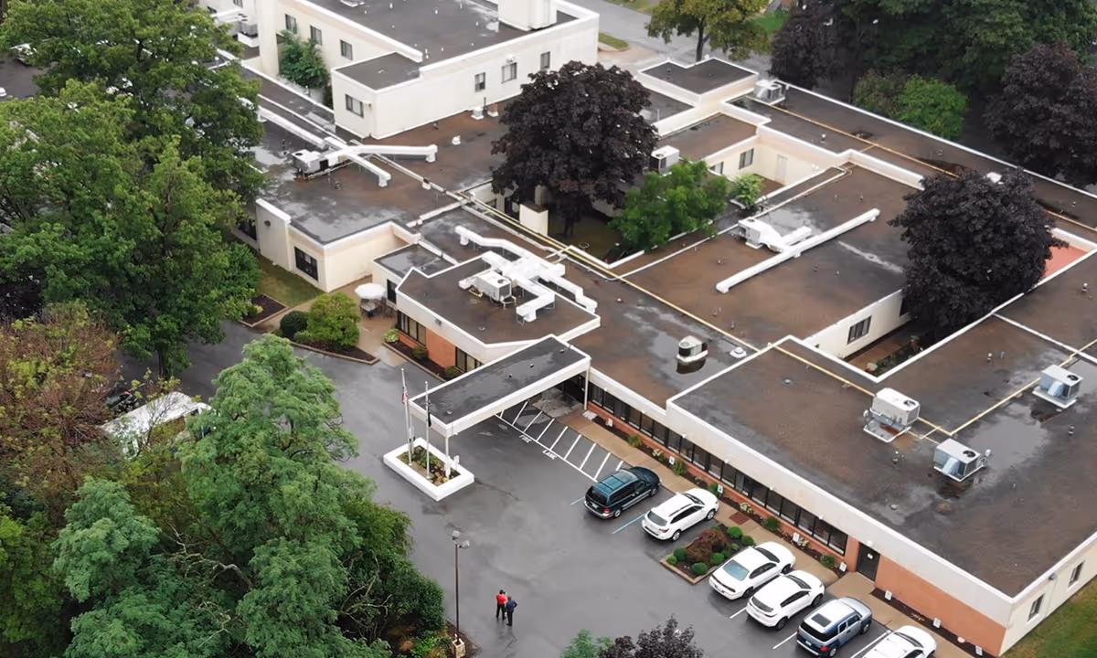 Aerial view of a single-story healthcare building showing its main entrance canopy, rooftop equipment, parked cars, and surrounding trees.