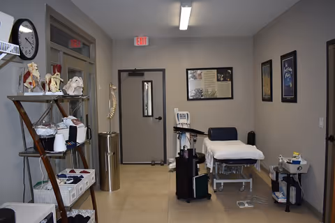 A medical examination room with a treatment table covered in white paper, medical equipment on a cart, anatomical models on a shelving unit, a clock on the wall, and framed posters. The room has beige walls and a door with a window and an exit sign above it.