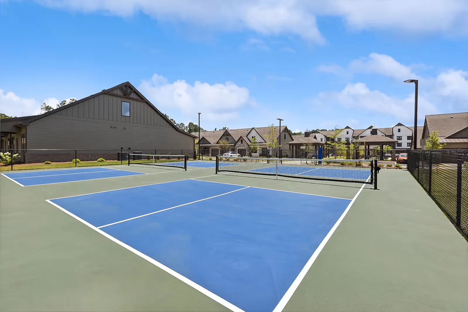 Outdoor view of a recreational area at The Phoenix at Opelika featuring multiple pickleball courts with blue and green surfaces, surrounded by a black chain-link fence. In the background, there are residential buildings with pitched roofs under a partly cloudy blue sky.