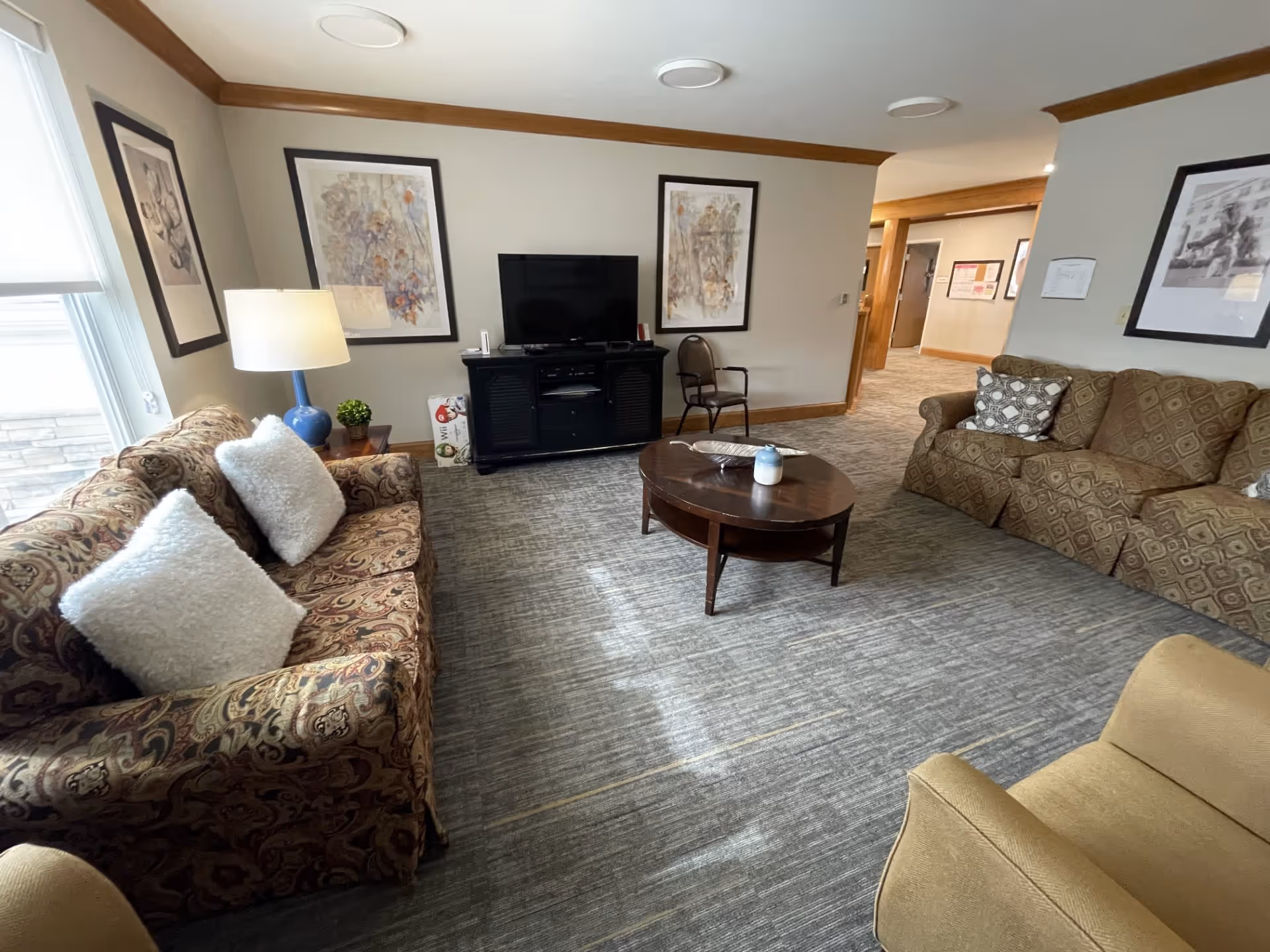 A cozy living room area with two patterned sofas, a beige armchair, a round wooden coffee table with a candle and decorative tray, a black TV stand with a flat-screen TV, a side table with a blue lamp and small plant, and framed artwork on the walls. The room has carpeted flooring and neutral-colored walls with wood trim.