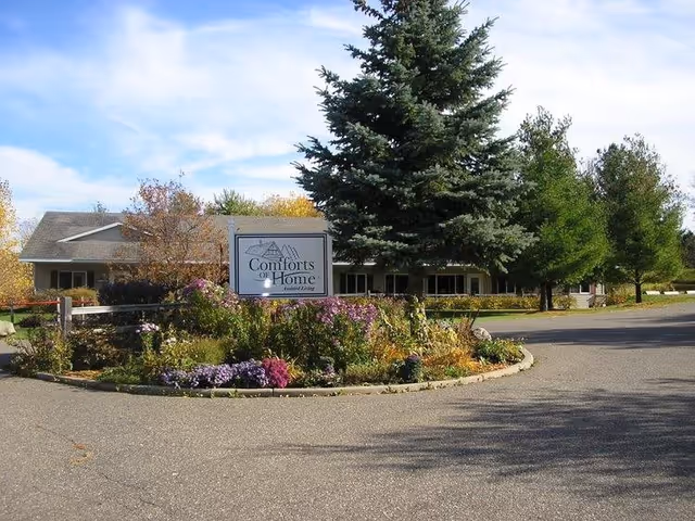 Exterior view of Comforts of Home Advanced Assisted Living and Men's Memory Care facility showing a driveway with a landscaped circular flower bed and a sign with the facility's name. Trees and a single-story building are visible in the background under a partly cloudy sky.