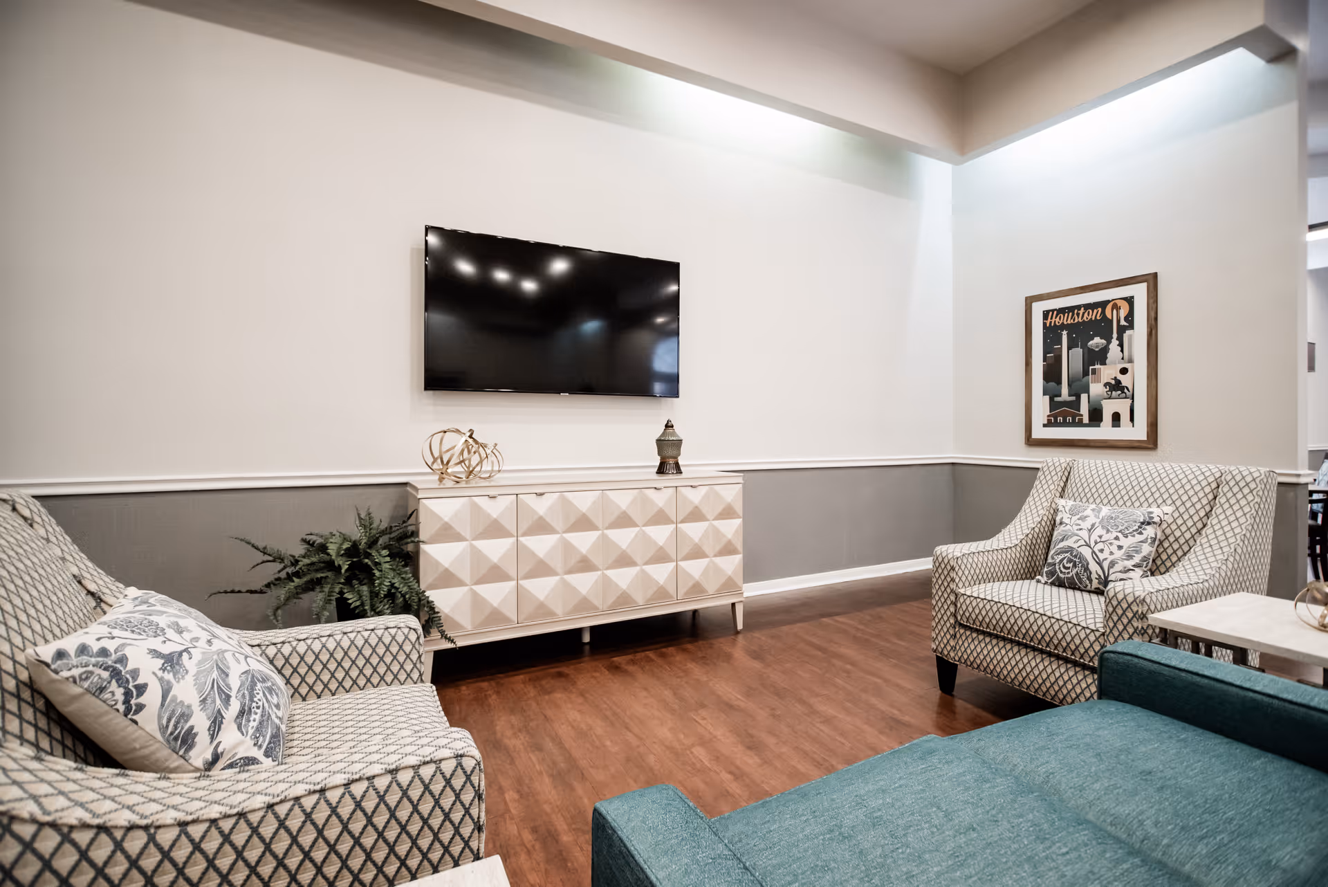 A cozy living room area with two patterned armchairs, a teal sofa, a white geometric cabinet, a flat-screen TV mounted on the wall, a framed Houston poster, and a potted plant on a wooden floor.