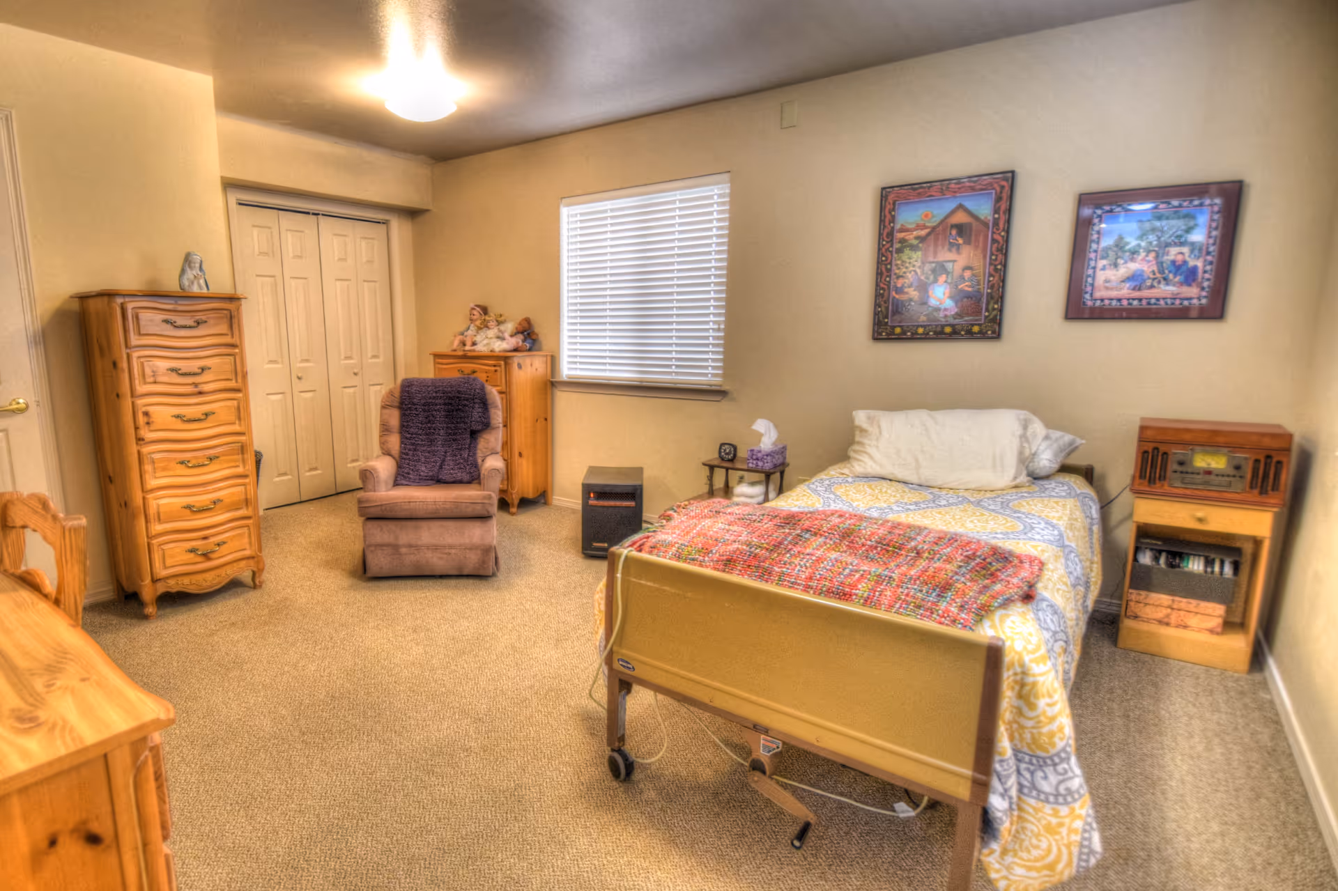 A cozy bedroom in a senior living facility featuring a single bed with a colorful blanket, a comfortable armchair with a purple throw, wooden dressers, a small side table with a tissue box, and two framed pictures on the wall. The room has beige walls and carpeted floor, with a window covered by blinds.