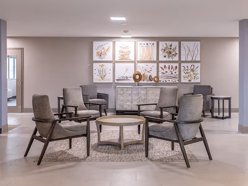 A modern sitting area in a senior living facility with six gray cushioned armchairs arranged around a round wooden coffee table on a textured rug. Behind the chairs is a decorative sideboard with abstract sculptures and a wall adorned with nine framed botanical prints. The room has neutral-colored walls and flooring, with a doorway visible on the left side.