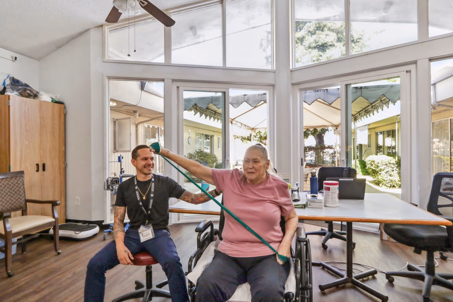A senior woman in a wheelchair exercises with a resistance band assisted by a male caregiver in a bright room with large windows and a ceiling fan. The room has wooden flooring, chairs, and a table with various items on it. Outside, there is a covered patio and greenery.