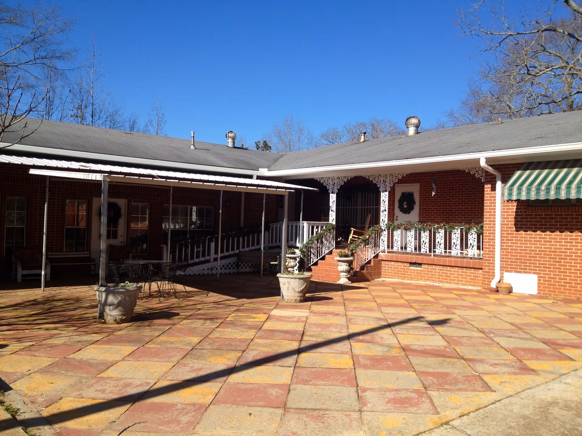 Outdoor courtyard area of a brick building with a tiled floor, white railings decorated with garlands, potted plants, a rocking chair on the porch, and a green and white striped awning. The sky is clear and blue.