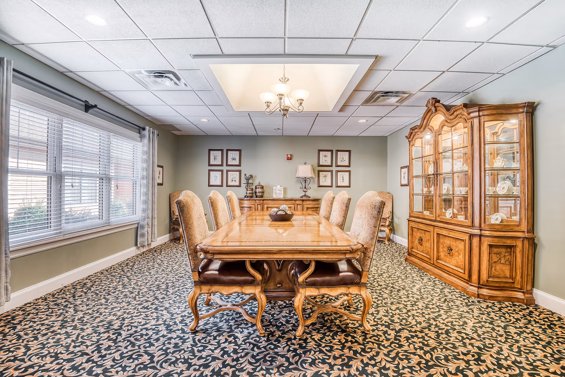 A formal dining room with a long wooden dining table surrounded by eight upholstered chairs. The room features a patterned carpet, a large window with blinds and curtains on the left, a chandelier hanging from a recessed ceiling, framed artwork on the walls, a wooden sideboard with decorative items, and a large wooden china cabinet displaying dishes on the right.