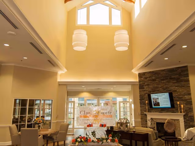 Bright and spacious senior living facility common area with high vaulted ceilings, large windows, and modern hanging light fixtures. The room features comfortable seating including armchairs and sofas arranged around a stone fireplace with a mounted TV. A table with flowers and a decorative sign is placed in the center, and large glass doors lead to an outdoor area.