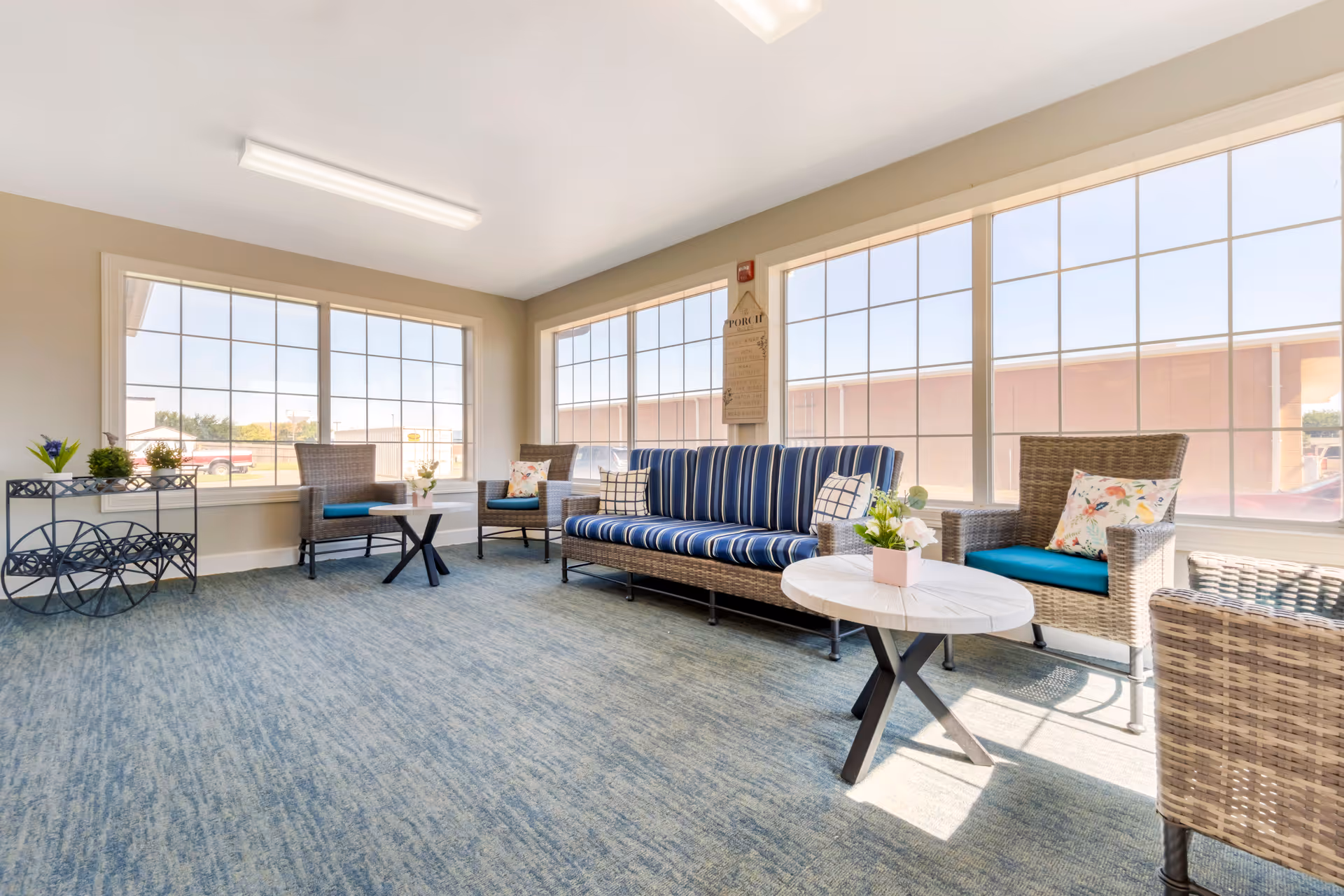 A bright and spacious sitting area with large windows letting in natural light. The room features a blue striped cushioned sofa, two wicker armchairs with blue cushions and floral pillows, a round white coffee table with a small flower arrangement, and a decorative black metal cart with plants. The carpet is blue, and the walls are painted beige.