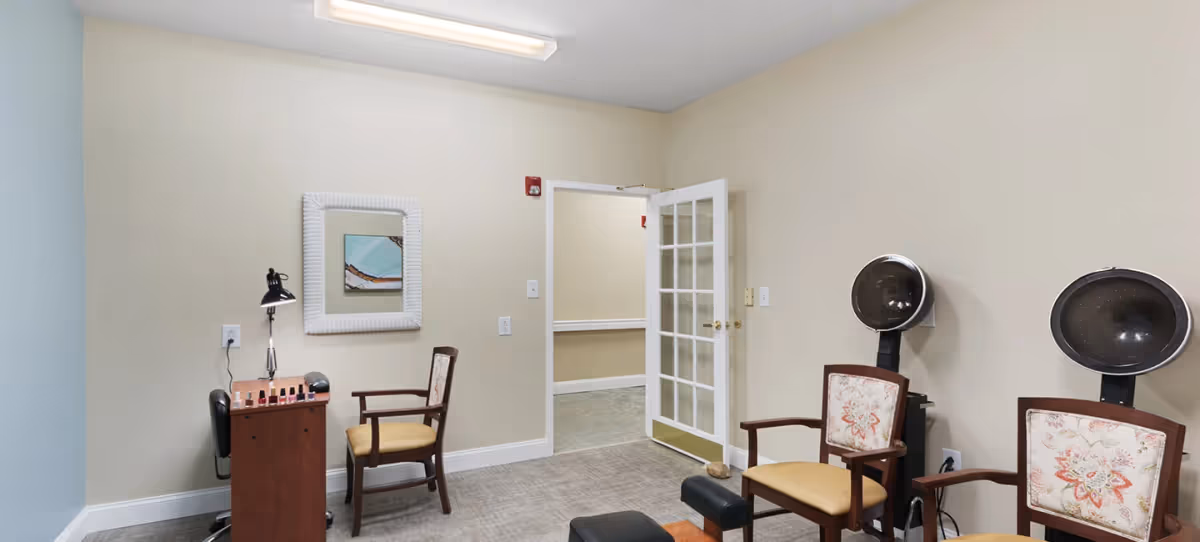 A small salon room with two vintage hair dryers, two wooden chairs with floral upholstery, a small wooden table with nail polish bottles and a desk lamp, a mirror on the wall, and a glass-paneled door leading to a hallway.