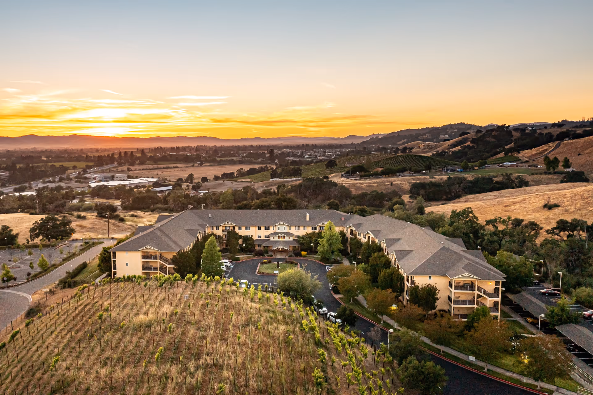 Aerial view of Solstice Senior Living at Santa Rosa facility at sunset, surrounded by rolling hills, vineyards, and trees with a clear sky and distant mountains in the background.