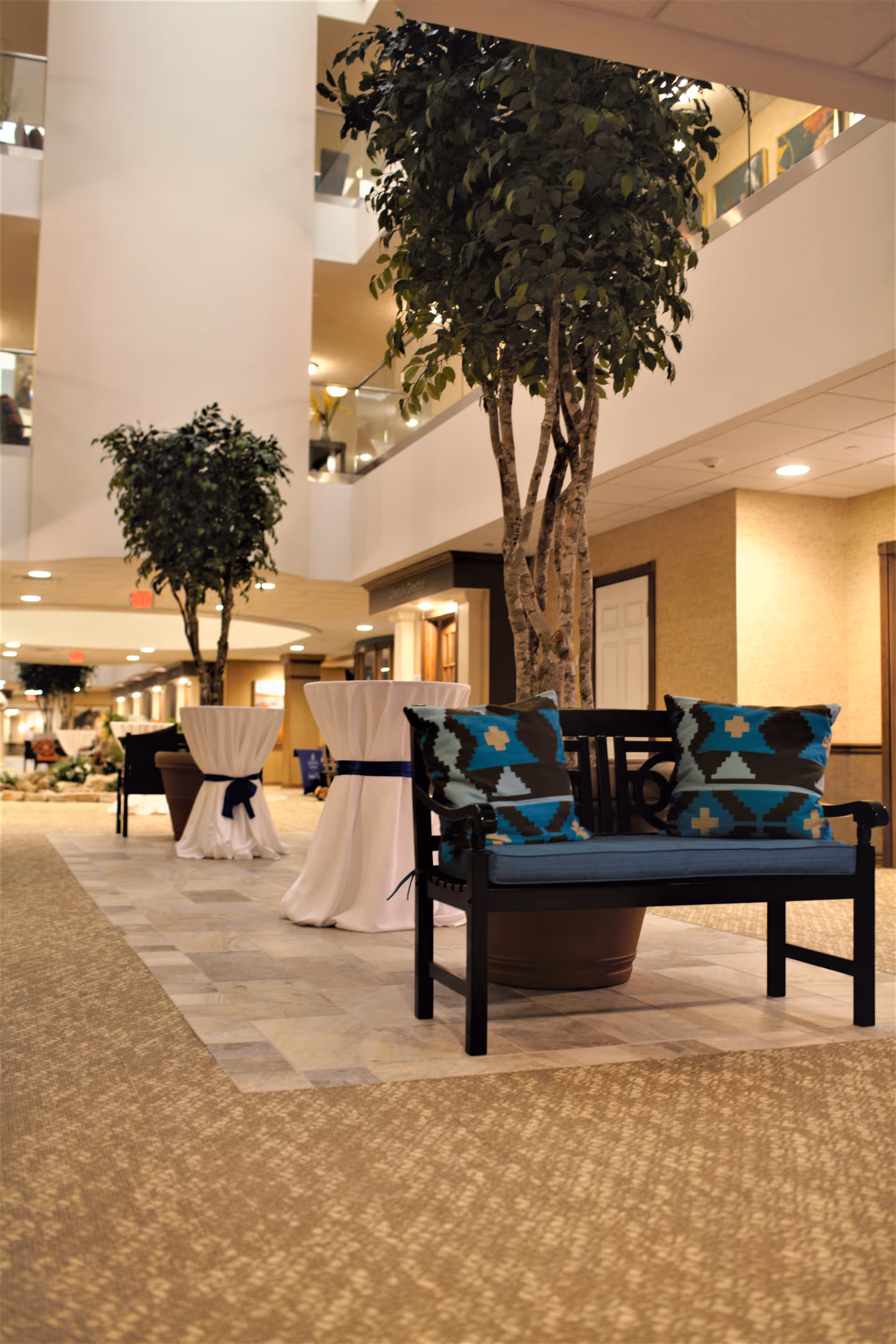 Indoor seating area in a senior living facility with a black bench featuring two blue patterned cushions, potted trees, and tall cocktail tables draped in white cloth with dark blue ribbons. The space has a beige carpet and tile flooring with a multi-level ceiling and balcony railings visible above.