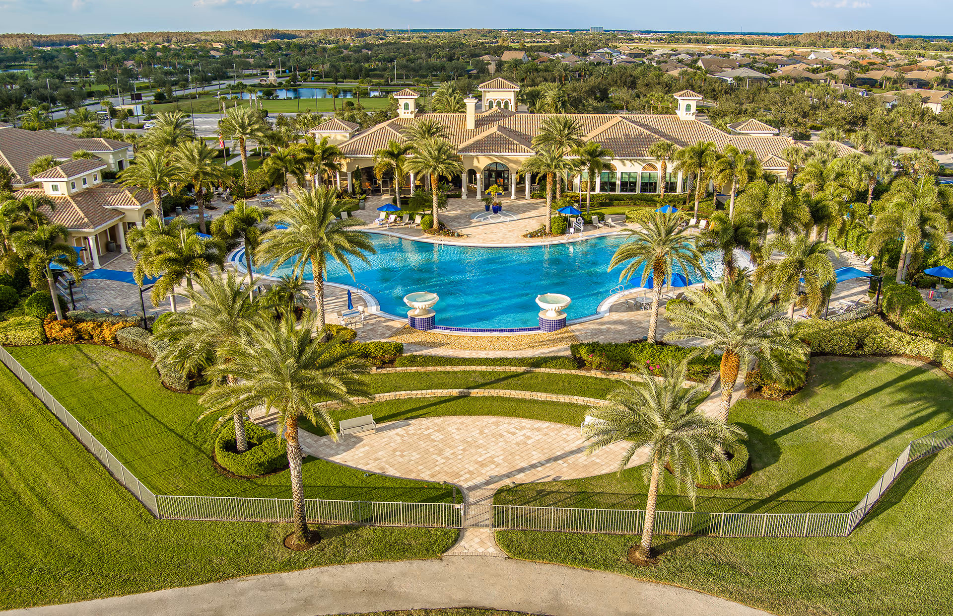 Aerial view of Del Webb Naples 55+ Retirement Community showing a large swimming pool surrounded by palm trees and a spacious clubhouse building with a tiled roof. The area is landscaped with green lawns, bushes, and paved walkways, with residential homes visible in the background.
