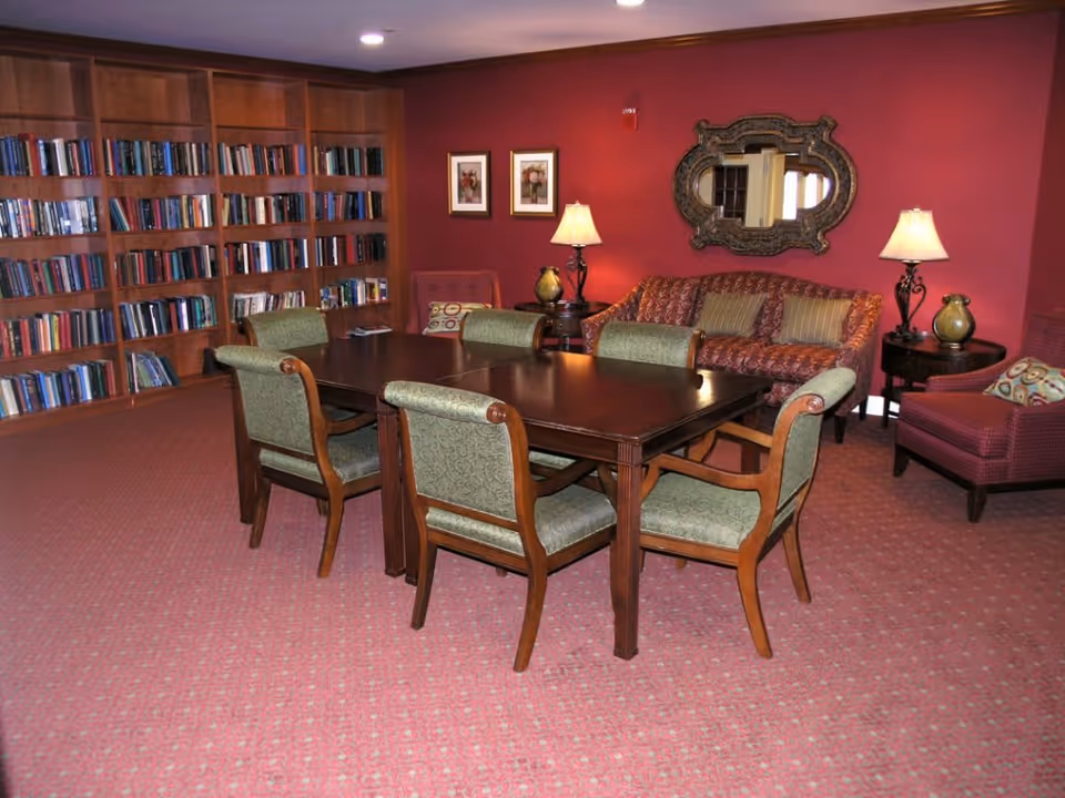 A cozy senior living common area with a dark wooden table surrounded by six green upholstered chairs. Behind the table is a red patterned sofa with striped cushions, two side tables with lamps, and a decorative mirror on a red wall. To the left, there is a large wooden bookshelf filled with books. The floor is carpeted in a red pattern.