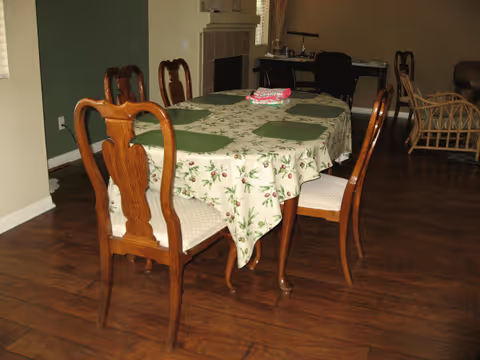 A dining room with a wooden table covered by a floral tablecloth and six wooden chairs with cushioned seats. The table has green placemats and a folded cloth in the center. The room has wooden flooring, a green accent wall, and a fireplace in the background. There is also a desk with a chair and a wicker chair visible in the background.