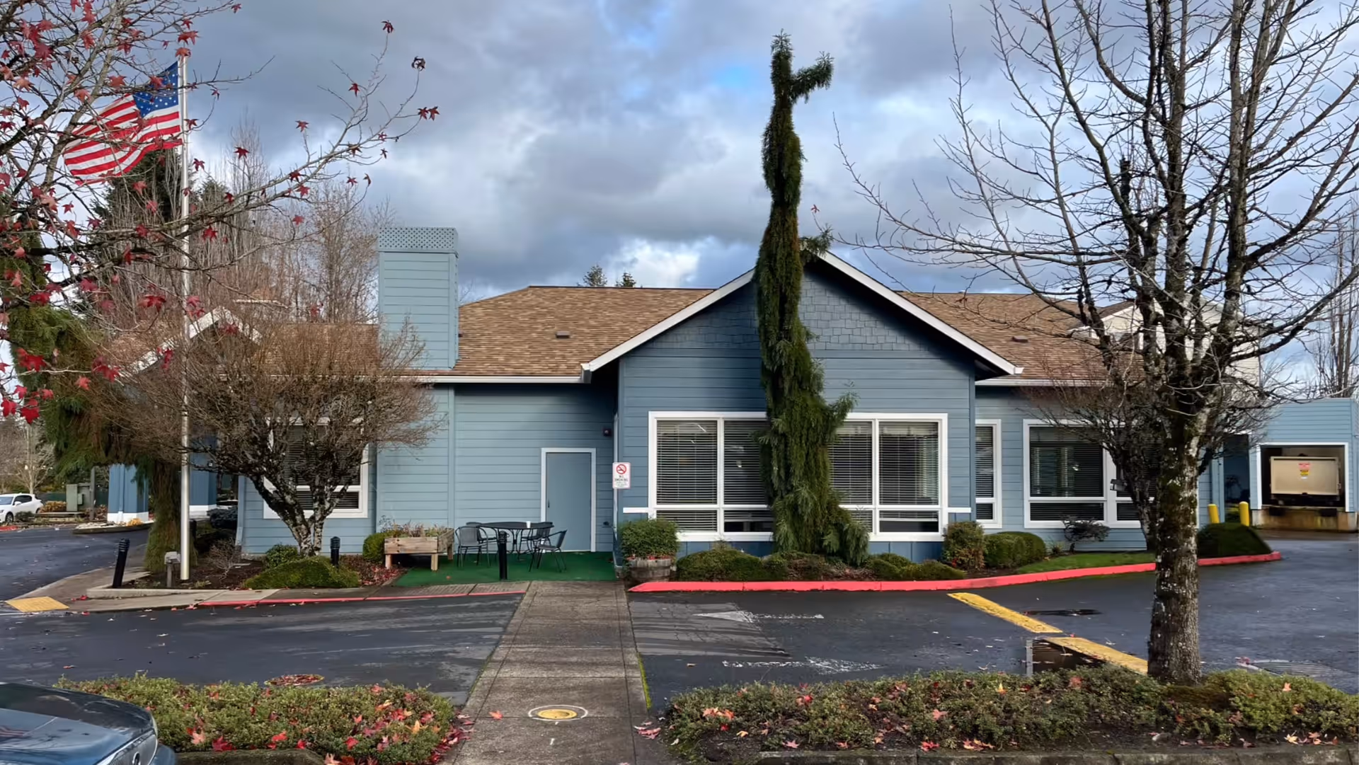 Blue, single-story senior living building with parking lot, trees, and an American flag.