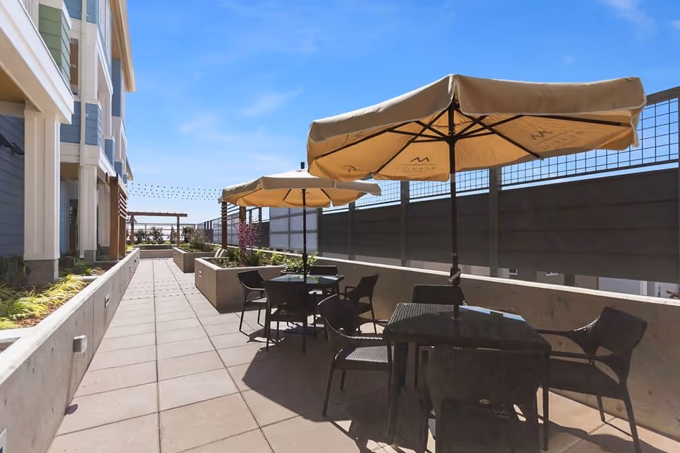 Outdoor patio area at Merrill Gardens at Rockridge with tables and chairs under large beige umbrellas, surrounded by concrete planters and a building on the left side under a clear blue sky.