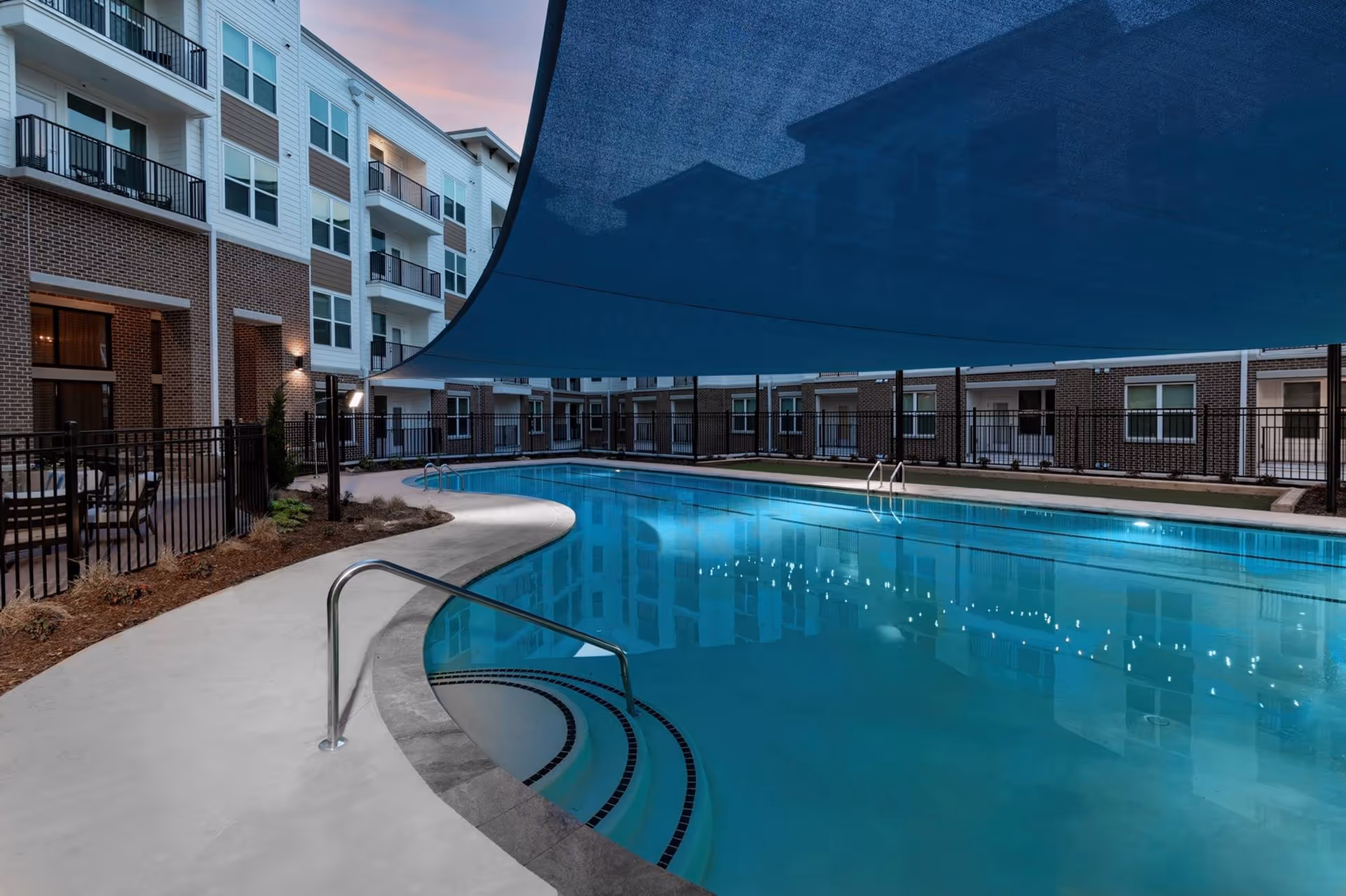 Outdoor swimming pool at a residential facility with a large blue shade canopy overhead, surrounded by a concrete deck and black metal fencing. The multi-story building with balconies and windows is visible in the background during dusk.