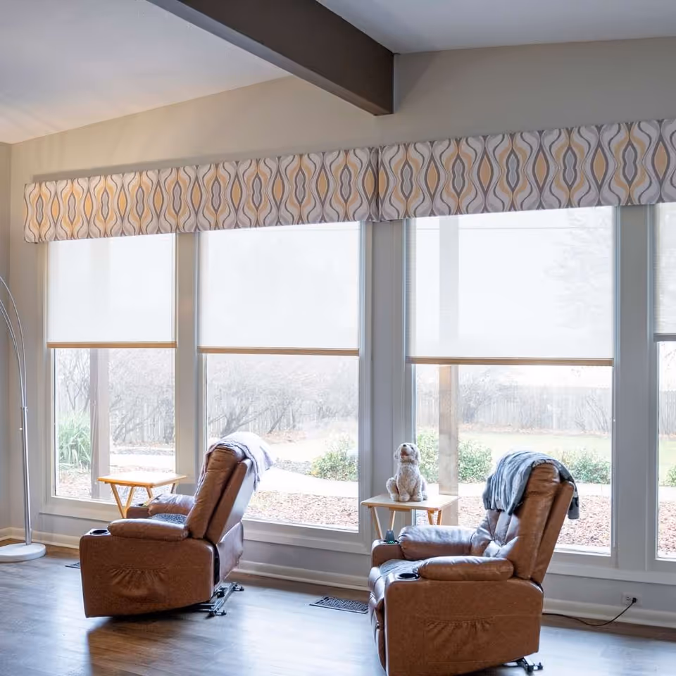 A bright sitting area with three tall windows and a patterned valance, featuring two brown recliners and small side tables (one with a stuffed dog).