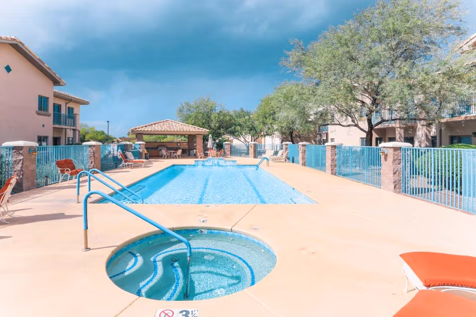 Outdoor swimming pool area at Mountain View Retirement Village featuring a rectangular pool with clear blue water, a circular hot tub in the foreground, lounge chairs with orange cushions, and a shaded pavilion in the background. The area is surrounded by a blue fence and trees, with multi-story residential buildings on either side.