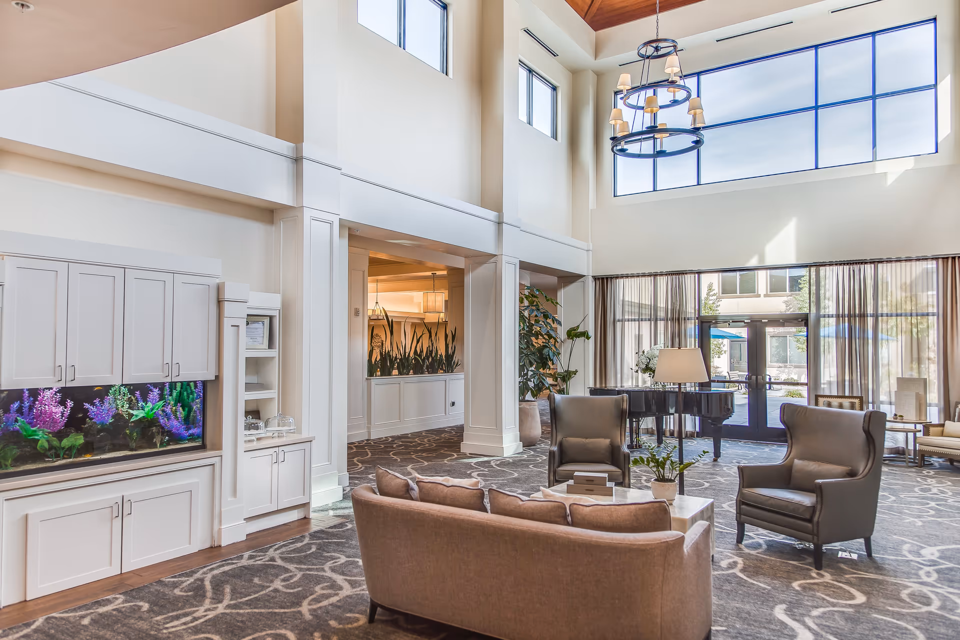 A spacious and well-lit living room area with high ceilings and large windows. The room features a beige sofa, two dark armchairs, a coffee table with a plant, a floor lamp, and a black grand piano near the glass doors leading outside. There is a built-in aquarium with colorful fish and plants on the left side, white cabinetry, and decorative plants throughout the space.