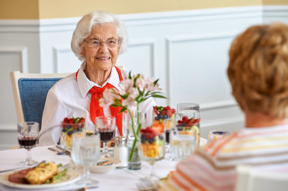 Two elderly women sitting at a dining table in a well-lit room, enjoying a meal with glasses of water, fruit cups, and plates of food. One woman with white hair and glasses is smiling and wearing a white shirt with a red scarf, while the other woman is seen from behind wearing a striped shirt. A vase with pink flowers is on the table.