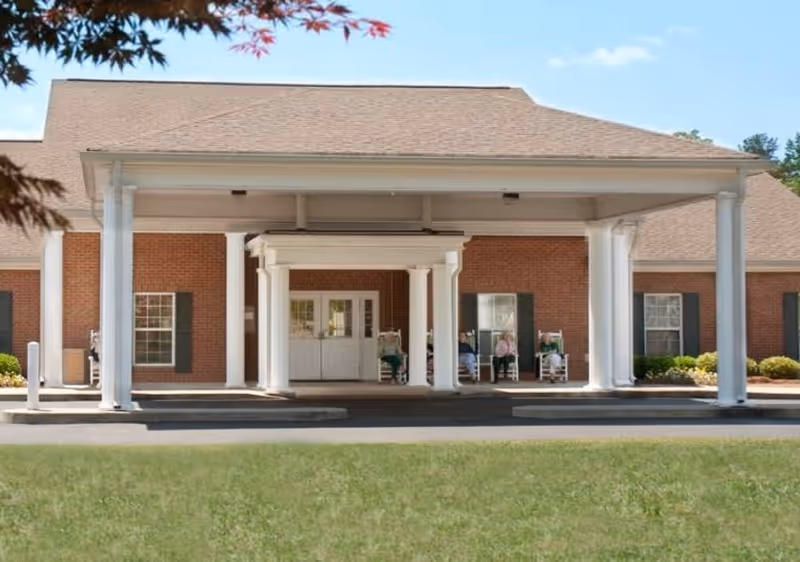 Front entrance of a brick building with white columns and a covered drop-off area. There are three elderly people sitting on rocking chairs on the porch near the entrance. The sky is clear and blue, and there is green grass in the foreground.