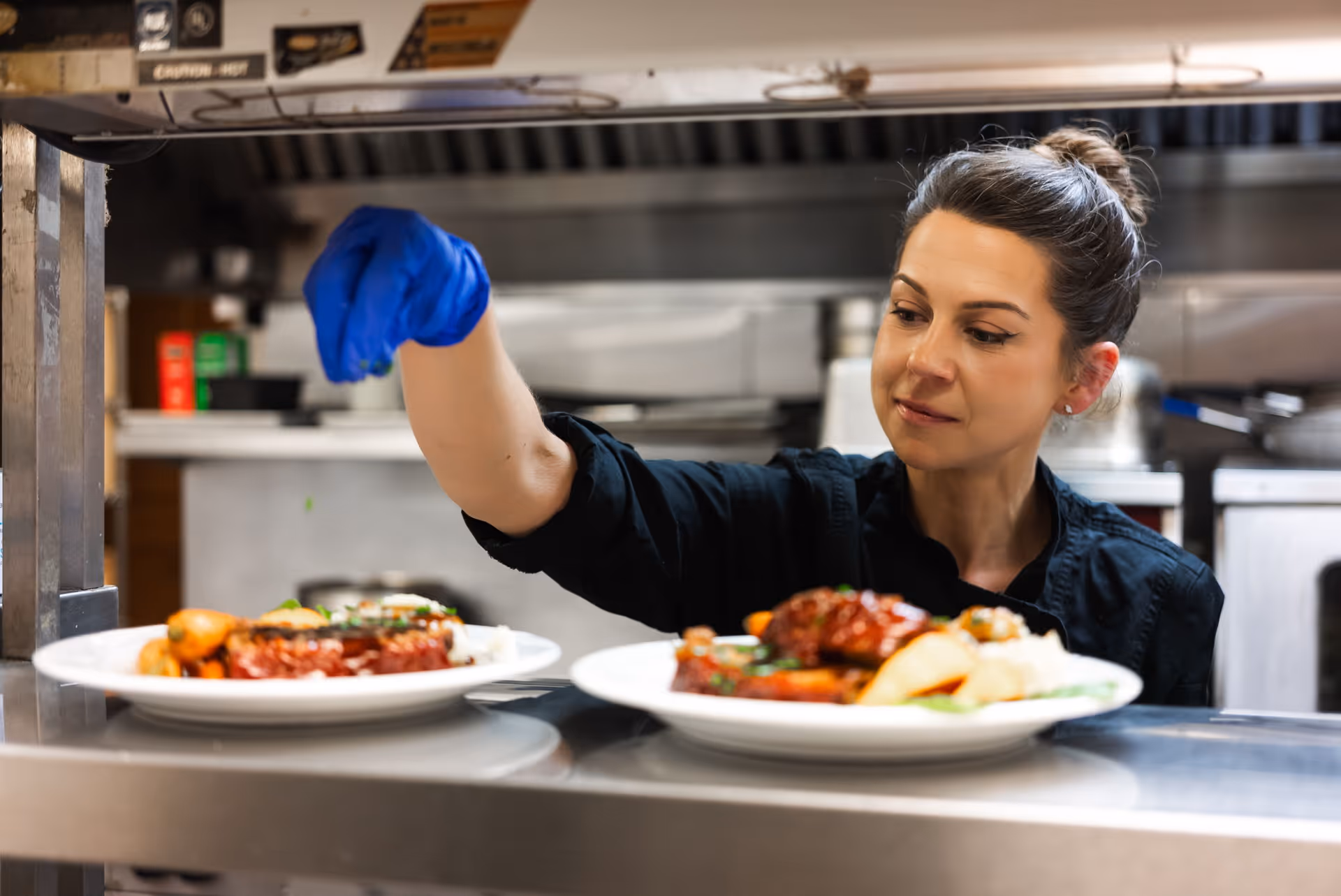 A woman wearing a black chef's uniform and a blue glove is placing plates of food on a serving counter in a commercial kitchen.