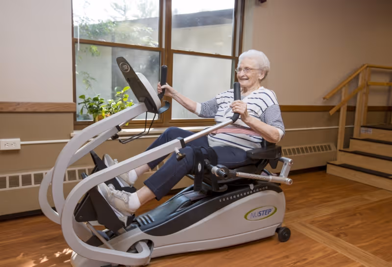 An elderly woman with white hair and glasses is smiling while using a NuStep seated exercise machine in a room with wooden floors, large windows, and a staircase in the background.