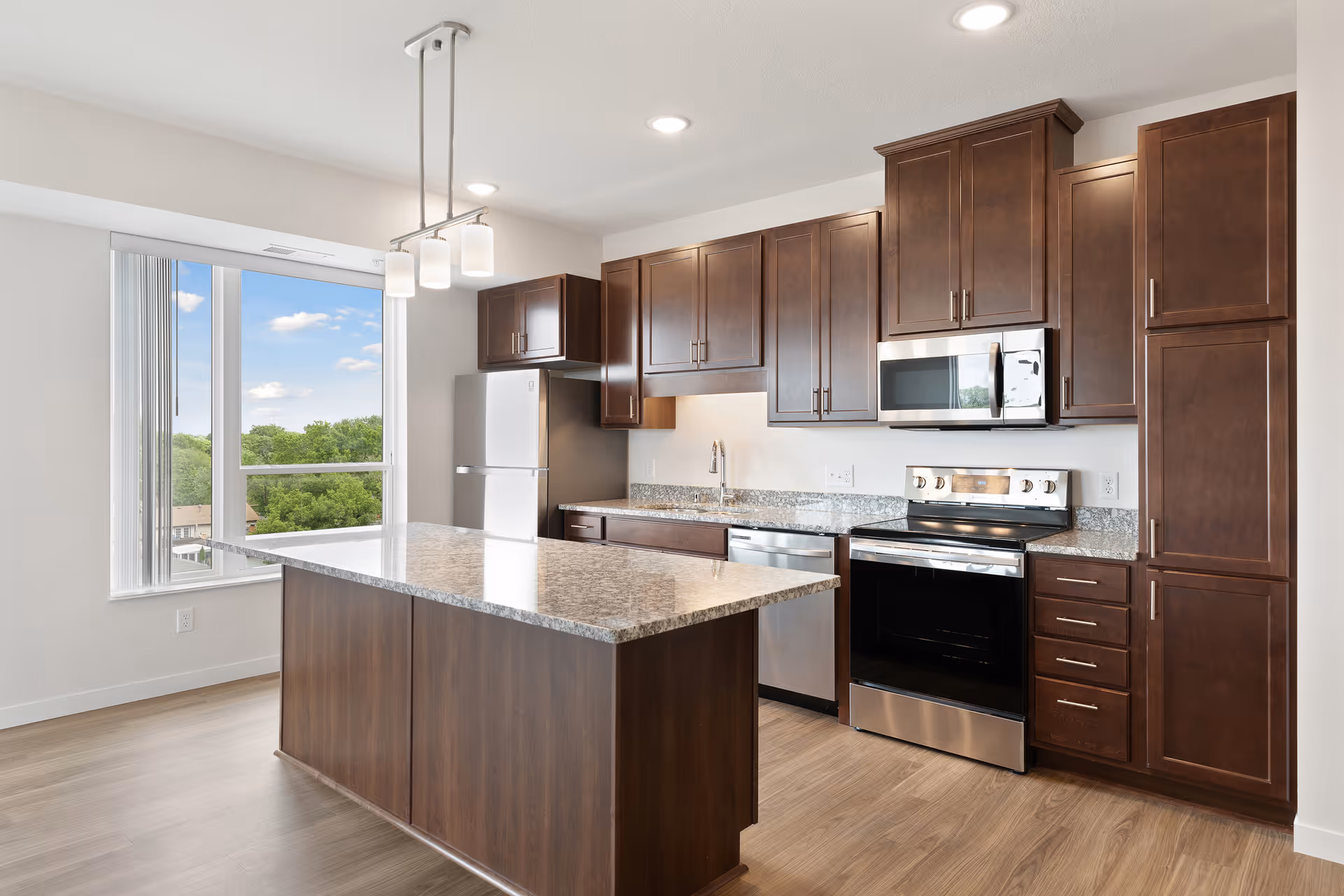 Modern kitchen with dark wood cabinets, stainless steel appliances including a refrigerator, oven, microwave, and dishwasher. There is a large island with a granite countertop in the center and a window showing a view of green trees and blue sky. The floor is wood and the ceiling has recessed lighting and a hanging light fixture above the island.