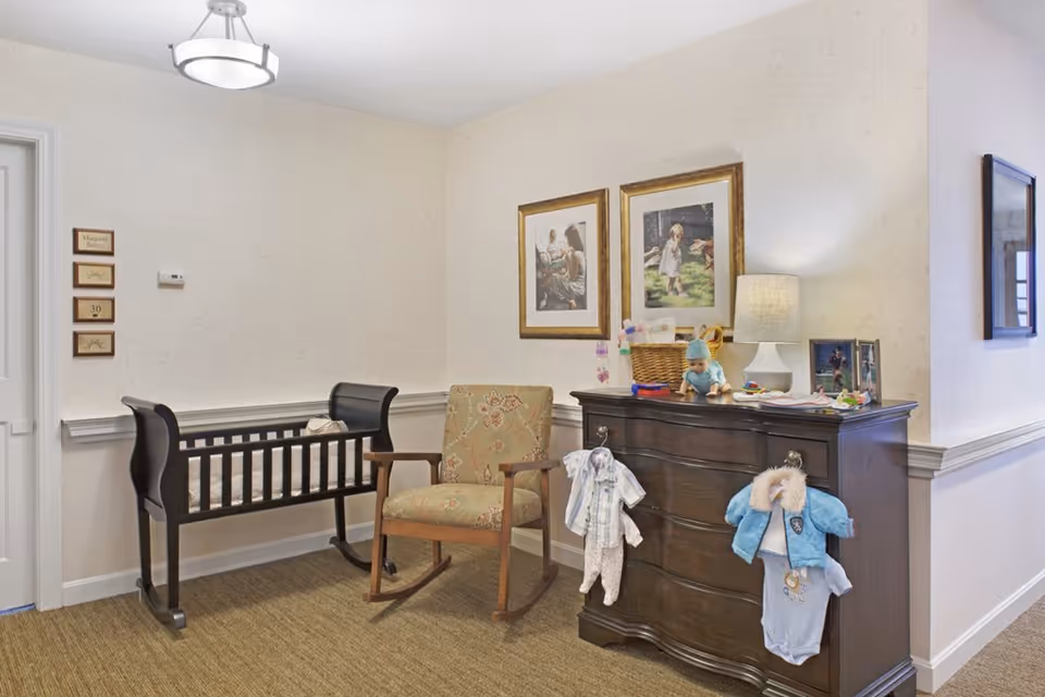 A cozy corner of a room featuring a dark wooden baby cradle, a cushioned rocking chair with floral upholstery, and a dark wooden dresser with baby clothes hanging on its handles. The dresser top holds a lamp, a basket, toys, and framed photos. Two framed pictures hang on the wall above the dresser. The room has light-colored wallpaper and carpeted flooring.