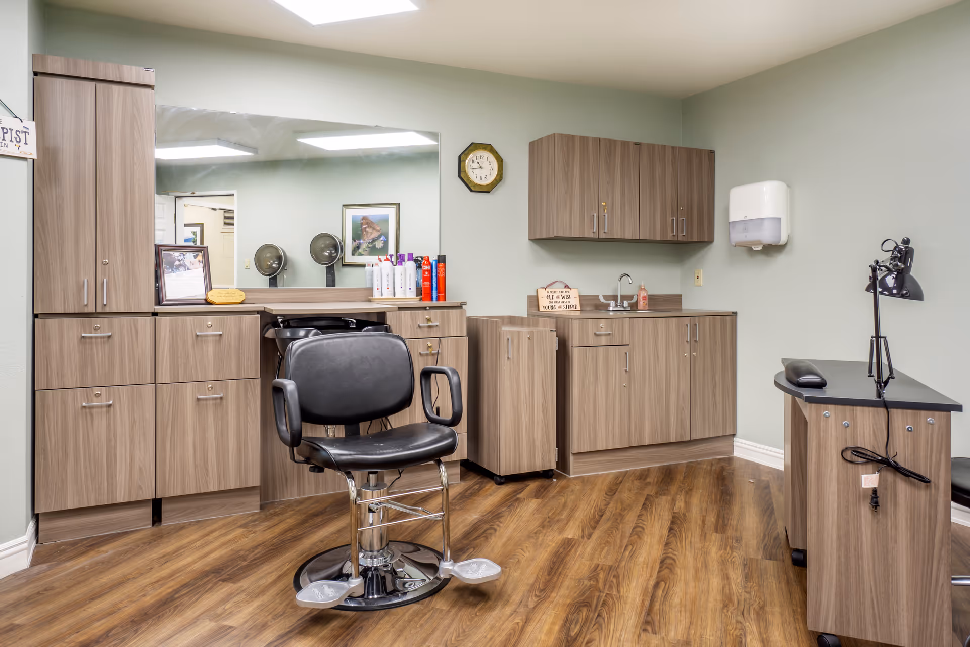 Interior salon room with a black barber chair in front of a large mirror, wood cabinets, a sink and hair care products.