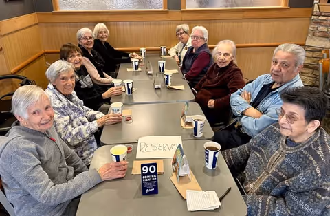 A group of elderly people sitting around a long table in a dining area, each holding or having a cup in front of them. The table has a reserved sign and a small card with the number 90. The room has wood-paneled walls and a stone accent on one side.