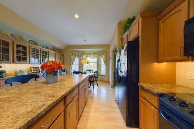 A bright kitchen with wooden cabinets and a large granite countertop island in the center. There are bar stools along one side of the island. The kitchen features a black refrigerator, a stove, and a microwave. In the background, there is a dining area with a table and chairs near a window with floral curtains and a hanging light fixture.