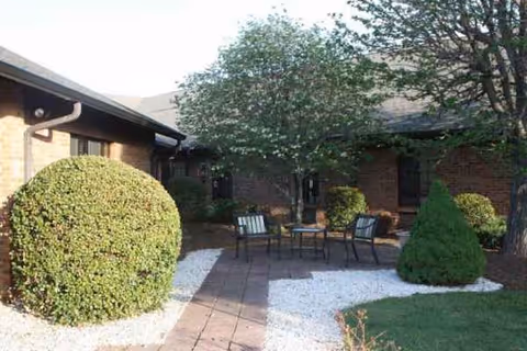 Outdoor courtyard with seating, bushes, trees, and a brick building in Margate Health and Rehab Center.