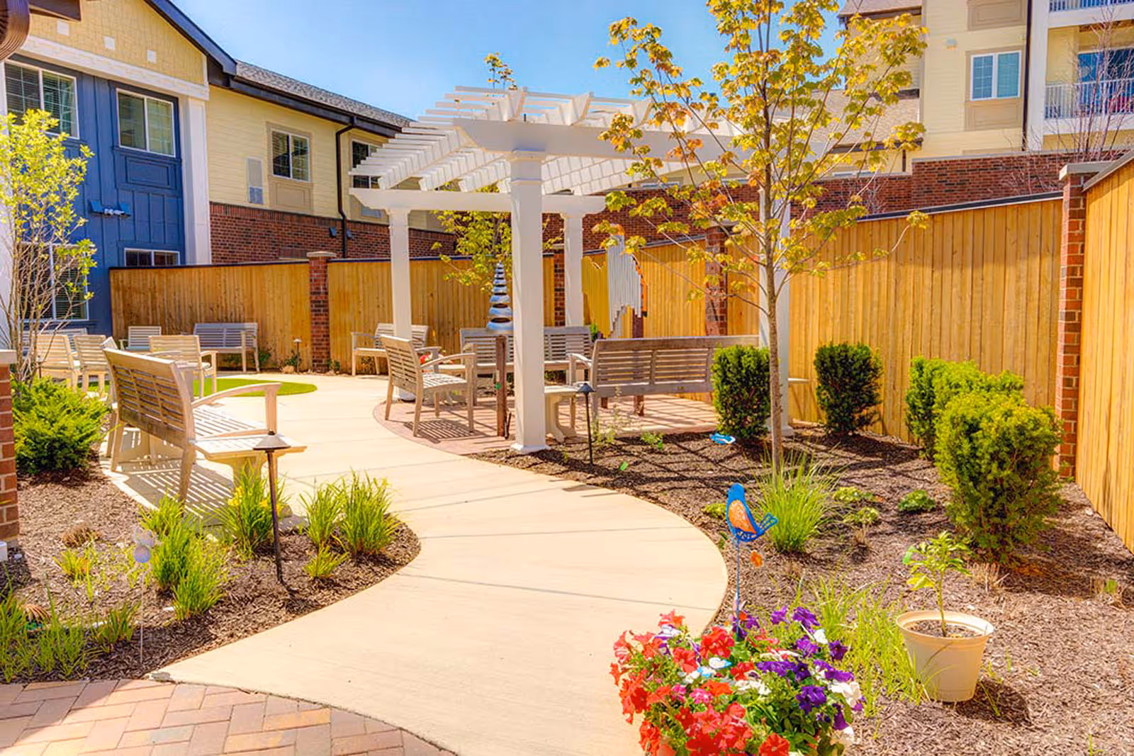 Sunny landscaped courtyard with a white pergola, benches, walkways, and potted flowers.