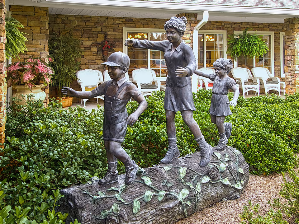 Bronze statue of three children balancing and walking on a large log surrounded by greenery, with a stone building and white wicker chairs on a porch in the background.