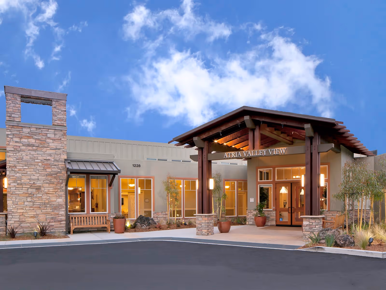 Front exterior view of Atria Valley View facility with a covered entrance supported by wooden beams, stone accents, large windows, benches, and potted plants under a partly cloudy blue sky.