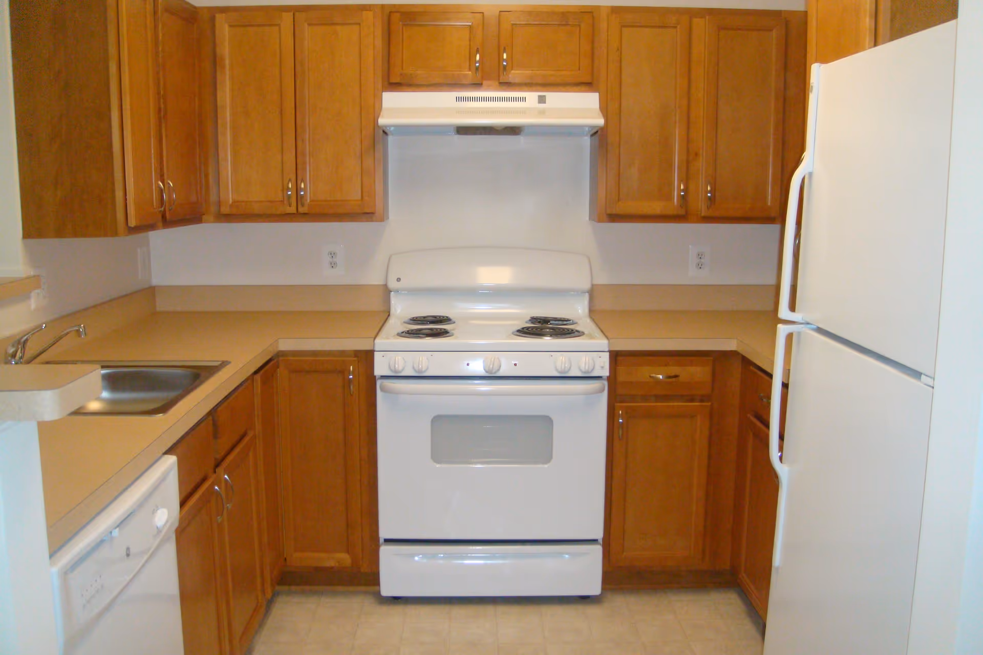A small kitchen with wooden cabinets, a white electric stove with four burners, a white refrigerator, a dishwasher, and a stainless steel sink. The countertops are beige, and the floor has light-colored tiles.