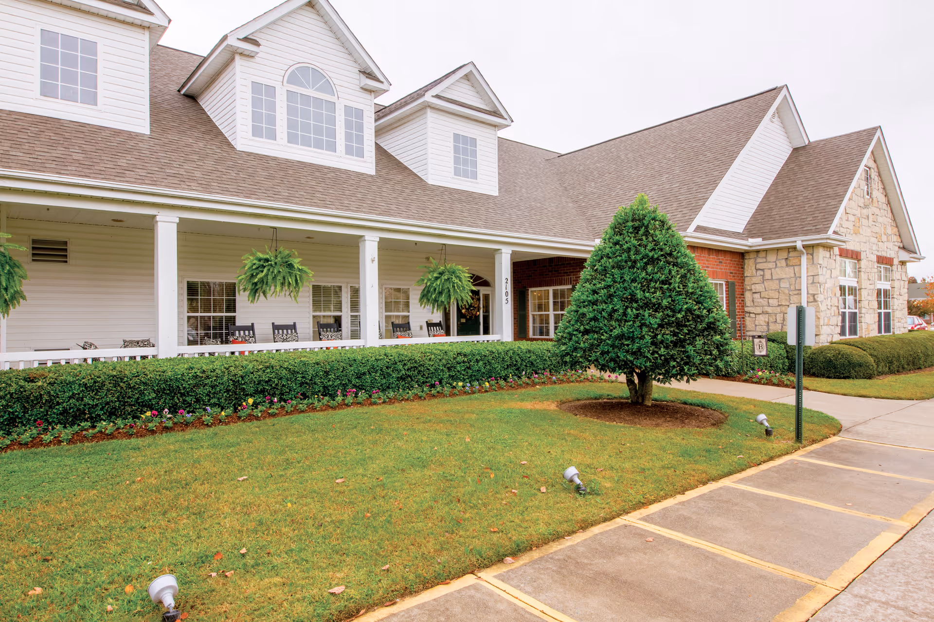 Exterior view of a senior living facility with a well-maintained lawn, a neatly trimmed tree, flower beds, and a covered porch with hanging plants and rocking chairs.
