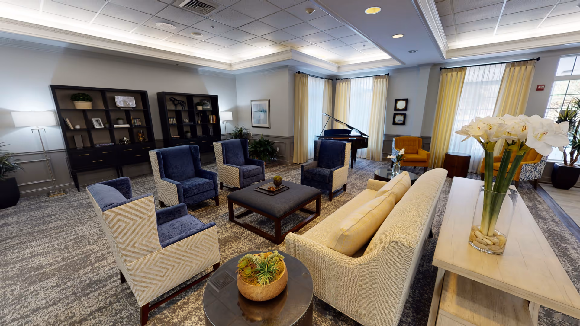 Bright common lounge with upholstered chairs and sofas arranged around a central ottoman, a grand piano by the windows, and decorative plants and flowers.