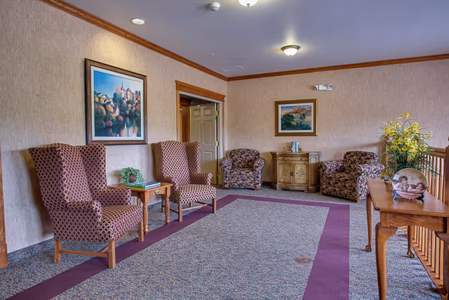 A cozy sitting area in a senior living facility with two patterned wingback chairs and a small wooden table on the left, two floral armchairs and a wooden cabinet with books in the background, and a wooden console table with a decorative plate and a vase of yellow flowers on the right. The walls are beige with framed landscape paintings, and the floor is carpeted with a patterned design and a purple border.