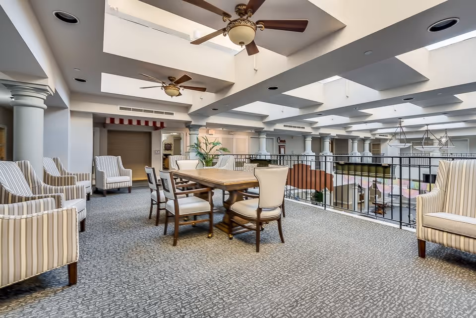 A spacious senior living common area with a high ceiling featuring multiple skylights and ceiling fans. The room has a carpeted floor, a wooden table surrounded by six chairs in the center, and several striped upholstered armchairs arranged along the walls. The area overlooks a lower floor through a black metal railing.