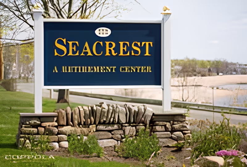 Blue and gold Seacrest entrance sign mounted on white posts over a stone base with grass, a road, and water in the background.