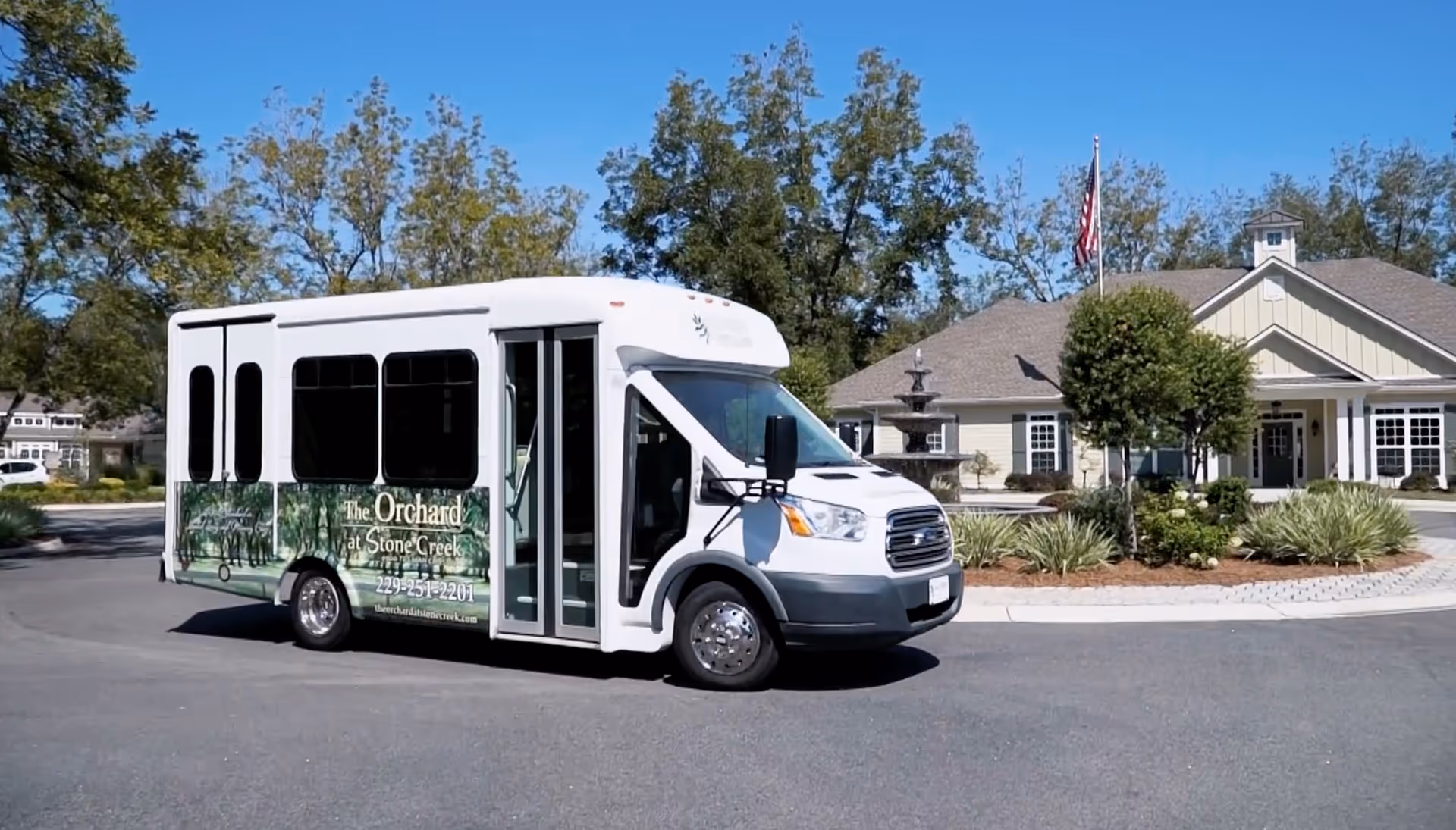 A white shuttle bus with green graphics and the text 'The Orchard at Stone Creek' parked on a circular driveway in front of a beige building with a fountain, trees, and an American flag on a sunny day.