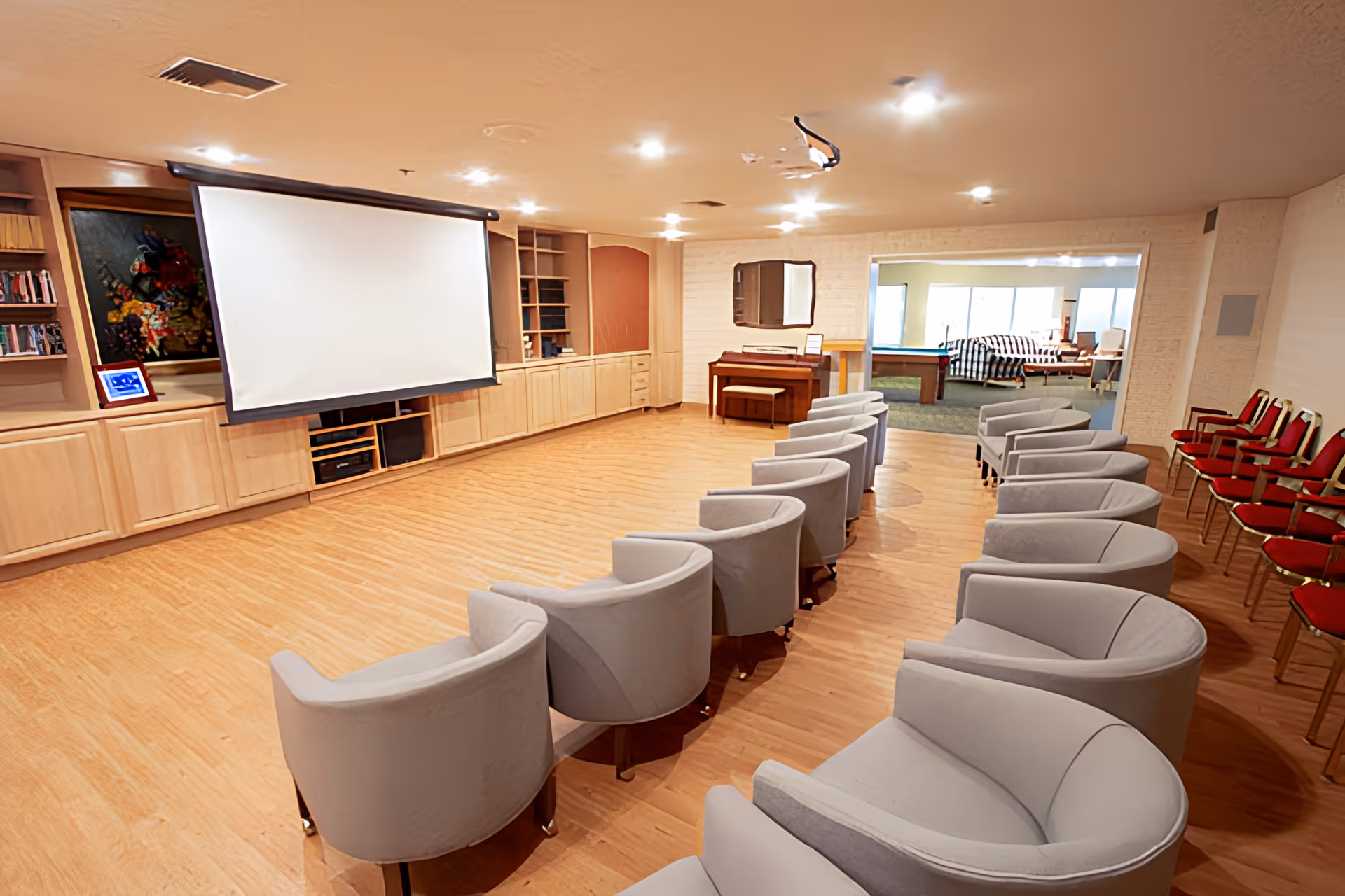 A room set up for presentations or movie viewing with a large white projection screen at the front, surrounded by built-in wooden cabinets and shelves. There are two rows of gray cushioned chairs arranged in a semi-circle facing the screen, and a row of red cushioned chairs along the right wall. In the background, there is an adjacent room with a pool table and seating area visible through a large open doorway.