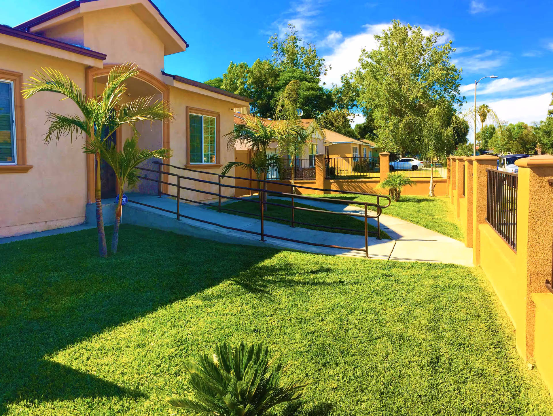 Exterior view of a beige building with a ramp leading to the entrance, surrounded by green grass, palm trees, and a yellow fence under a blue sky with some clouds.